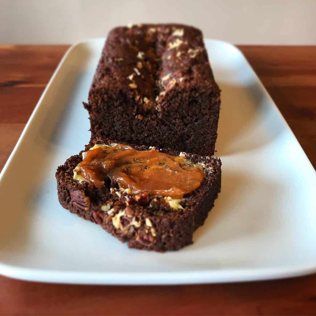 Pecan Molasses Bread on a white platter with one slice spread with butter and sweet potato butter, all on a wooden table