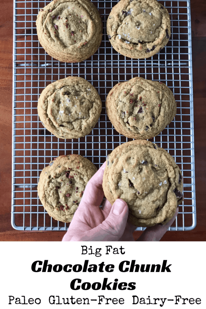 A hand holding a Big Fat Chocolate Chunk Cookie with the other cookies on a cooling rack on a wooden table