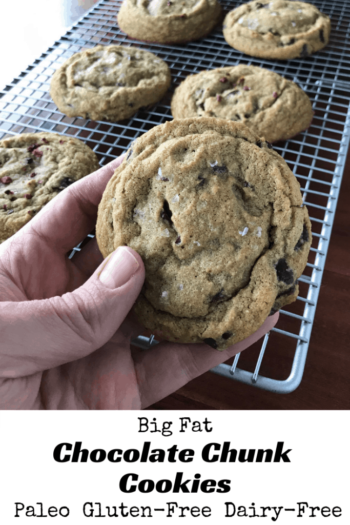 A hand holding a Big Fat Chocolate Chunk Cookie with other cookies on a cooling rack on a wooden table