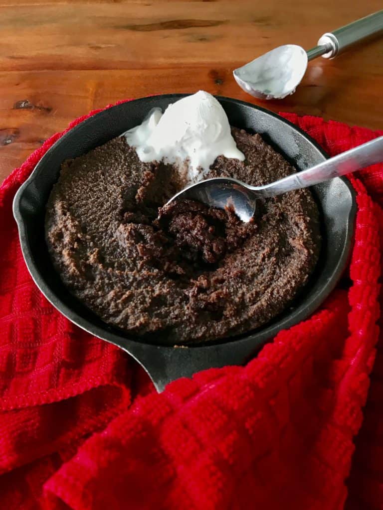 Chocolate cake in a cast iron pan wrapped in a red towel, topped with ice cream with a spoon digging in and a messy ice scream scoop on the table in the background