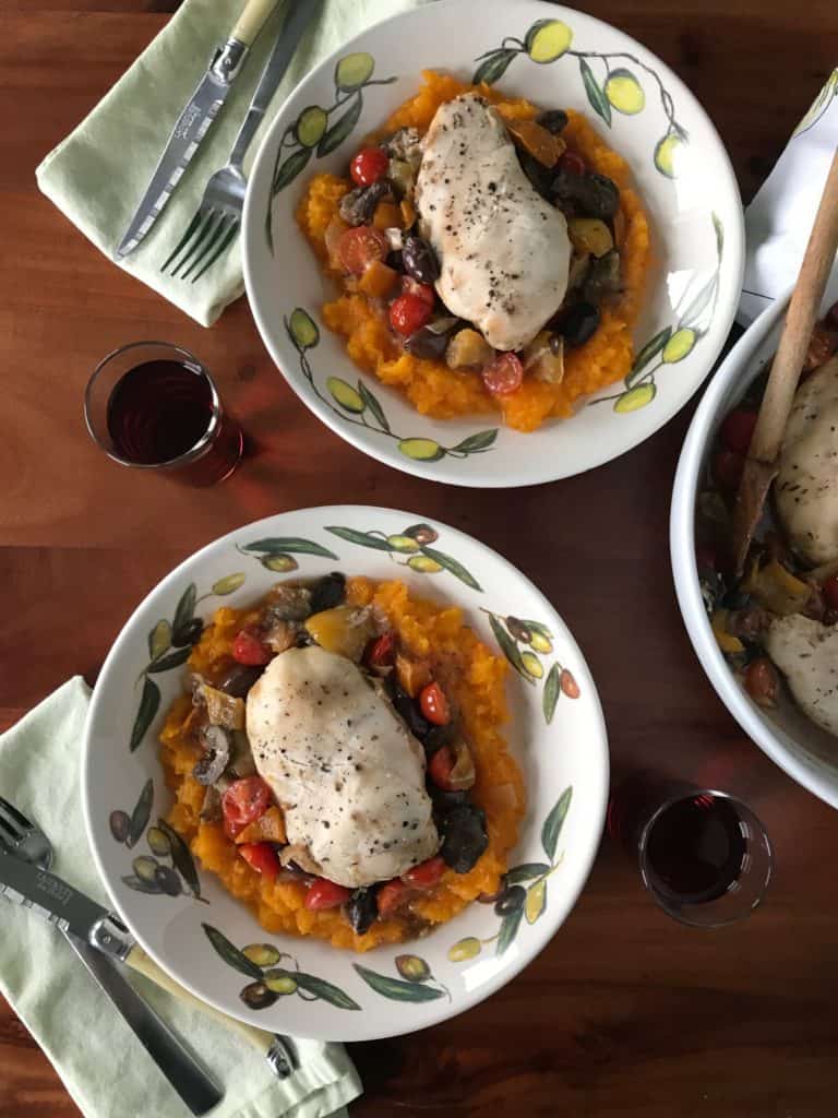Slow-Cooker Italian Chicken Stew on mashed butternut squash in white bowls surrounded by glasses of wine, napkins, silverware and a white serving bowl with a wood handle, all on a wood table