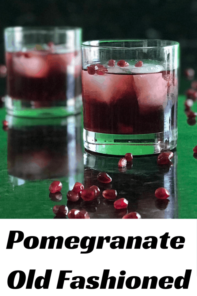 2 Pomegranate Old Fashioneds in rocks glasses on black marble surrounded by pomegranate arils