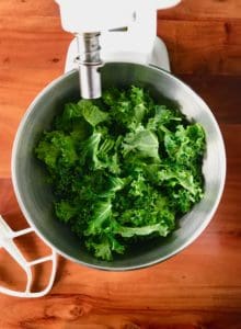 Raw kale leaves in the metal bowl of a white stand mixer with the white paddle attachment laying next to it on a wooden table