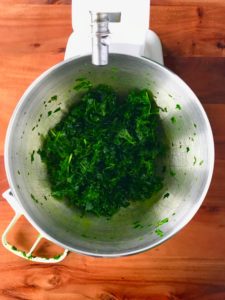 Tenderized kale leaves in the metal bowl of a white stand mixer with the used white paddle attachment laying next to it on a wooden table