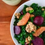 Kale Salad with Granola, Avocado Green Goddess Dressing and a Secret in a white bowl next to a glass pitcher of dressing on a wooden table
