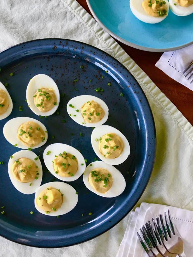 A blue platter of Caesar Deviled Eggs next to a light blue plate with more eggs surrounded by napkins and forks