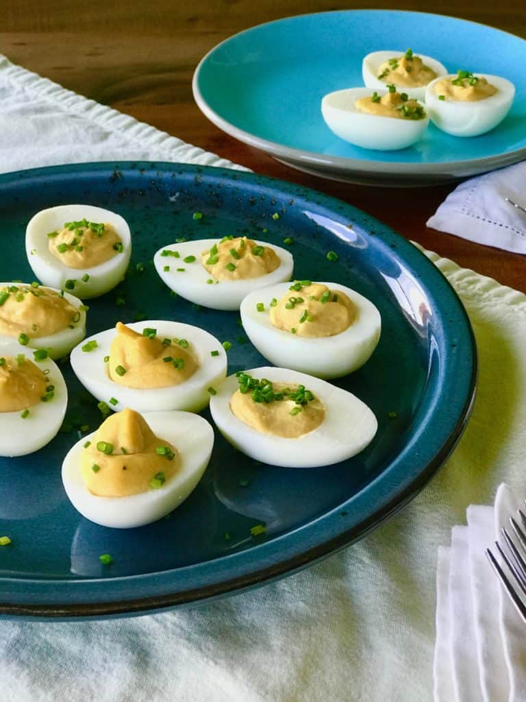 A blue platter of Caesar Deviled Eggs next to a light blue plate with more eggs surrounded by napkins and forks
