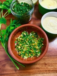 A close-up of gremolata in a wooden bowl on a wooden table next to parsley and other condiments