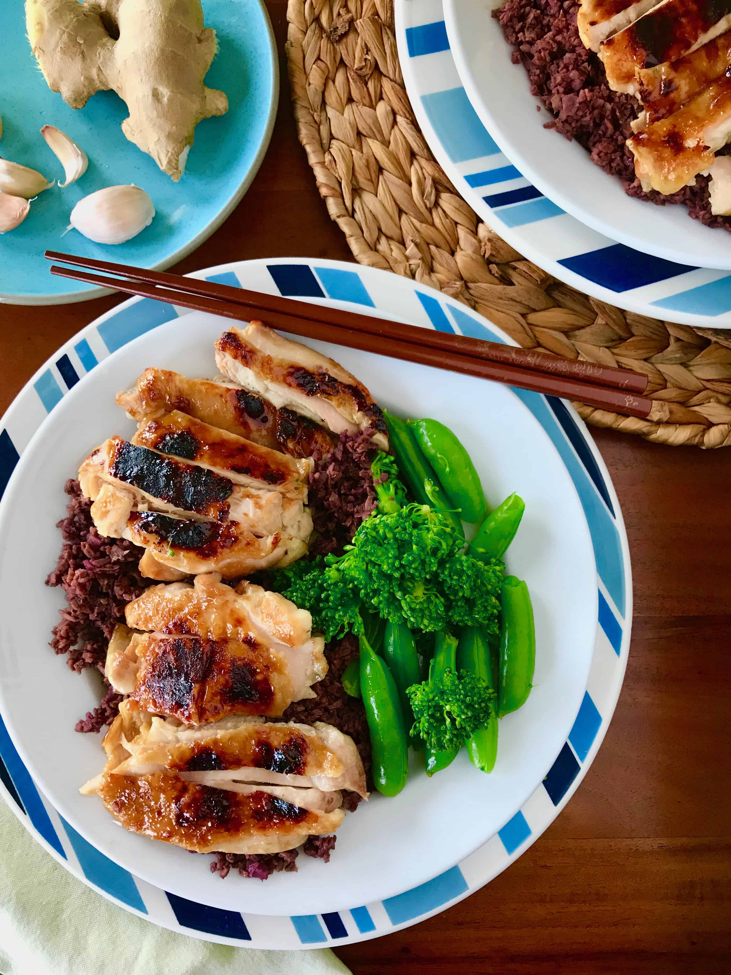 Healthy Teriyaki Chicken on two white plates over blue striped plates with a green napkin and chopsticks, next to a blue plate of ginger and garlic, all on top of a wooden table