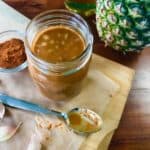 A jar of Tequila Pineapple Barbecue Sauce on a cutting board surrounded by a spoon, garlic, a bowl of cayenne, a pineapple and a bottle of tequila on a wooden table