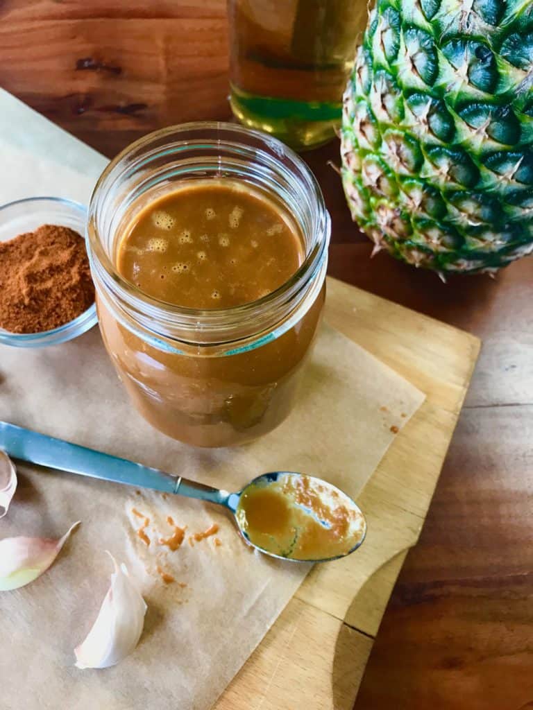 A jar of Tequila Pineapple Barbecue Sauce on a cutting board surrounded by a spoon, garlic, a bowl of cayenne, a pineapple and a bottle of tequila on a wooden table