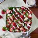 Strawberry, Cucumber and Arugula Salad with Poppy Seed Yogurt Dressing in a white square bowl on a green napkin on a wooden table next to a serving glass of dressing
