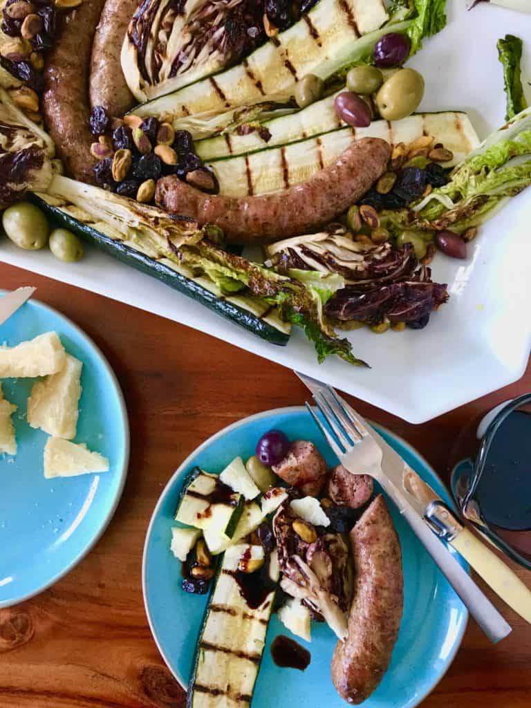 Grilled Italian Platter with Italian sausage, zucchini, romaine lettuce and raddichio with pistachios, dried cherries and olives on a white platter on a wooden table with Parmesan on the side