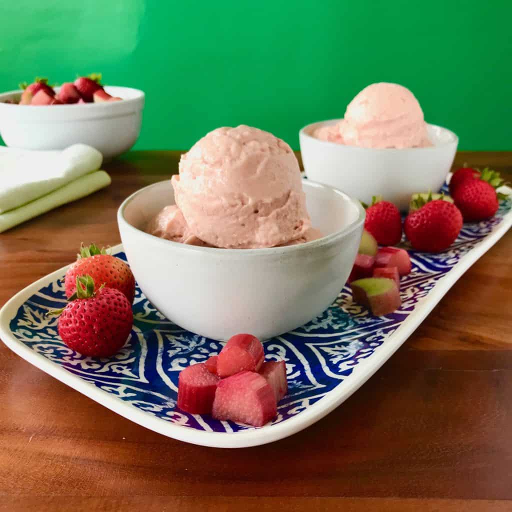 Dairy-Free Rhubarb Gelato with Easy Strawberry Sauce in a white bowl on a blue platter with strawberries and rhubarb pieces with a white bowl and green napkins in the background, all on a wooden table