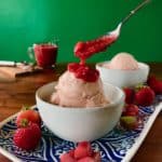 Dairy-Free Rhubarb Gelato with Easy Strawberry Sauce in a white bowl on a blue platter with strawberries and rhubarb pieces with a glass pitcher full of the sauce and an ice cream scoop in the background, all on a wooden table