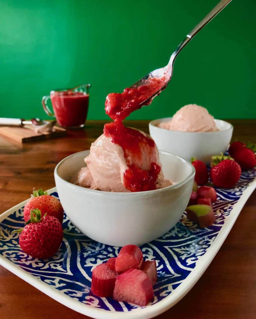 Dairy-Free Rhubarb Gelato with Easy Strawberry Sauce in a white bowl on a blue platter with strawberries and rhubarb pieces with a glass pitcher full of the sauce and an ice cream scoop in the background, all on a wooden table
