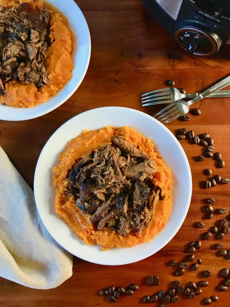 Slow Cooker Espresso Pulled Pork on mashed sweet potatoes on white plates by coffee beans, all on a wooden table