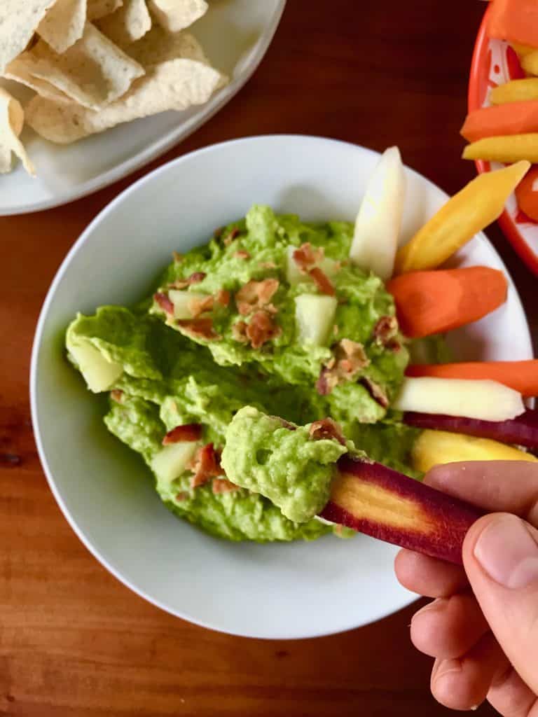 Bacon Pineapple Guacamole in a white bowl on a wooden table served with carrots and chips