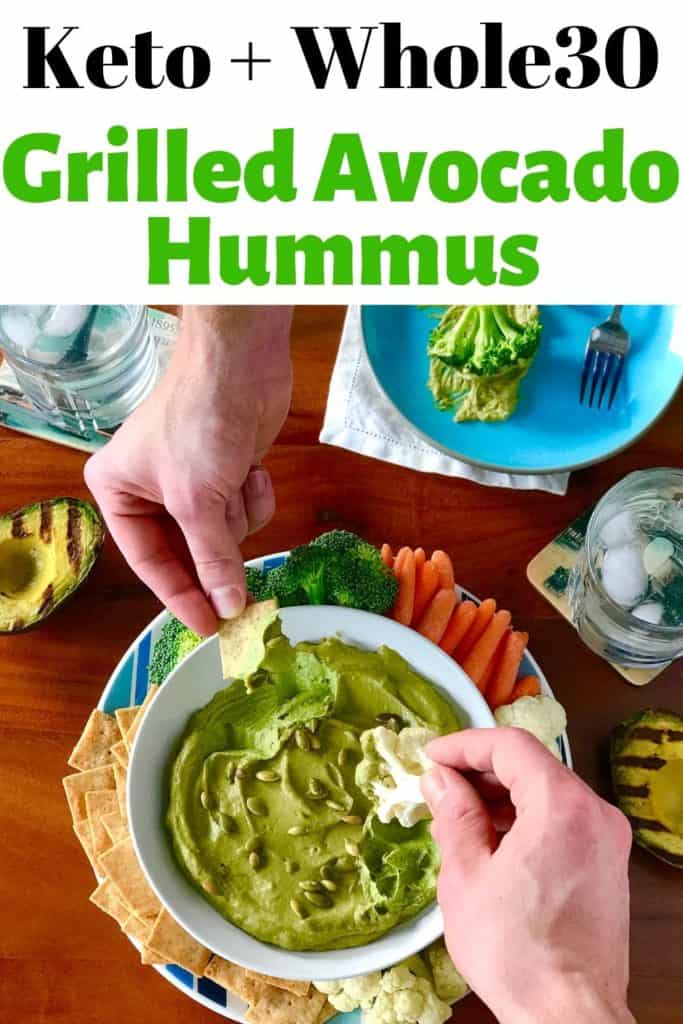 Grilled Avocado Hummus in a white bowl, with hands dipping in crackers and veggies, surrounded by grilled avocados, all on a wooden table