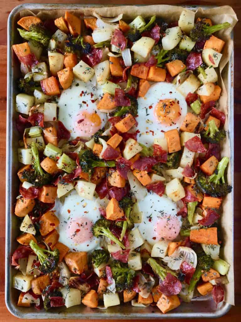 Sheet Pan Breakfast Hash on a baking sheet with potatoes, sweet potatoes, broccoli, onions, salami and eggs, all on a wooden table