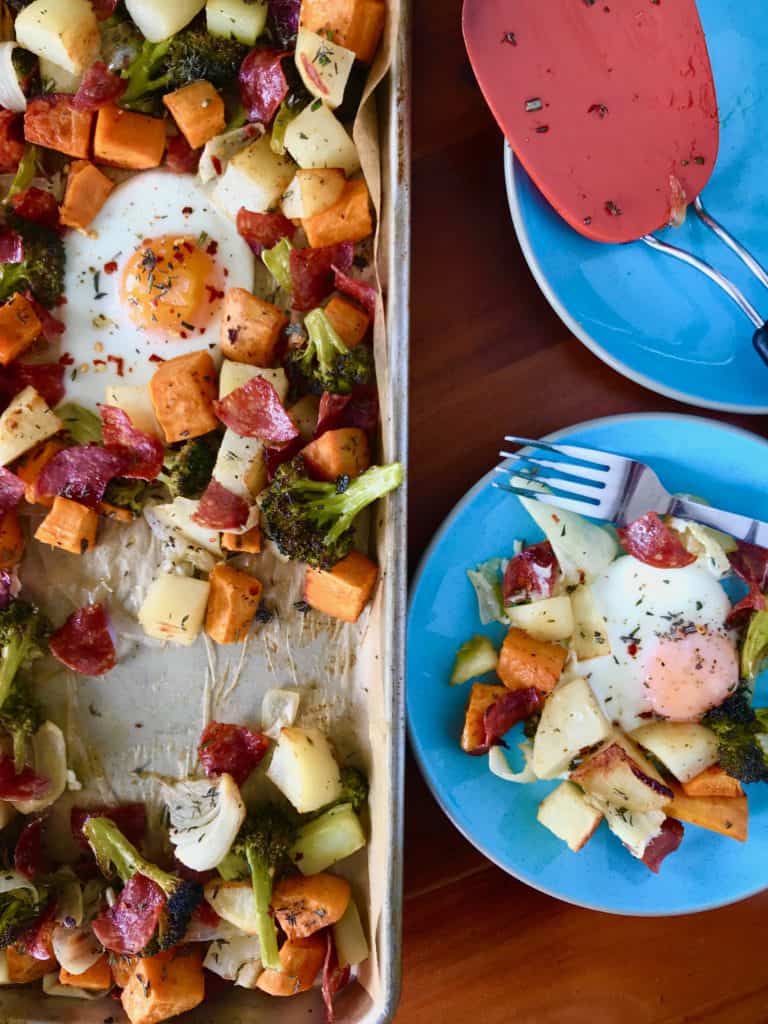 Sheet Pan Breakfast Hash on a baking sheet with potatoes, sweet potatoes, broccoli, onions, salami and eggs, all on a wooden table