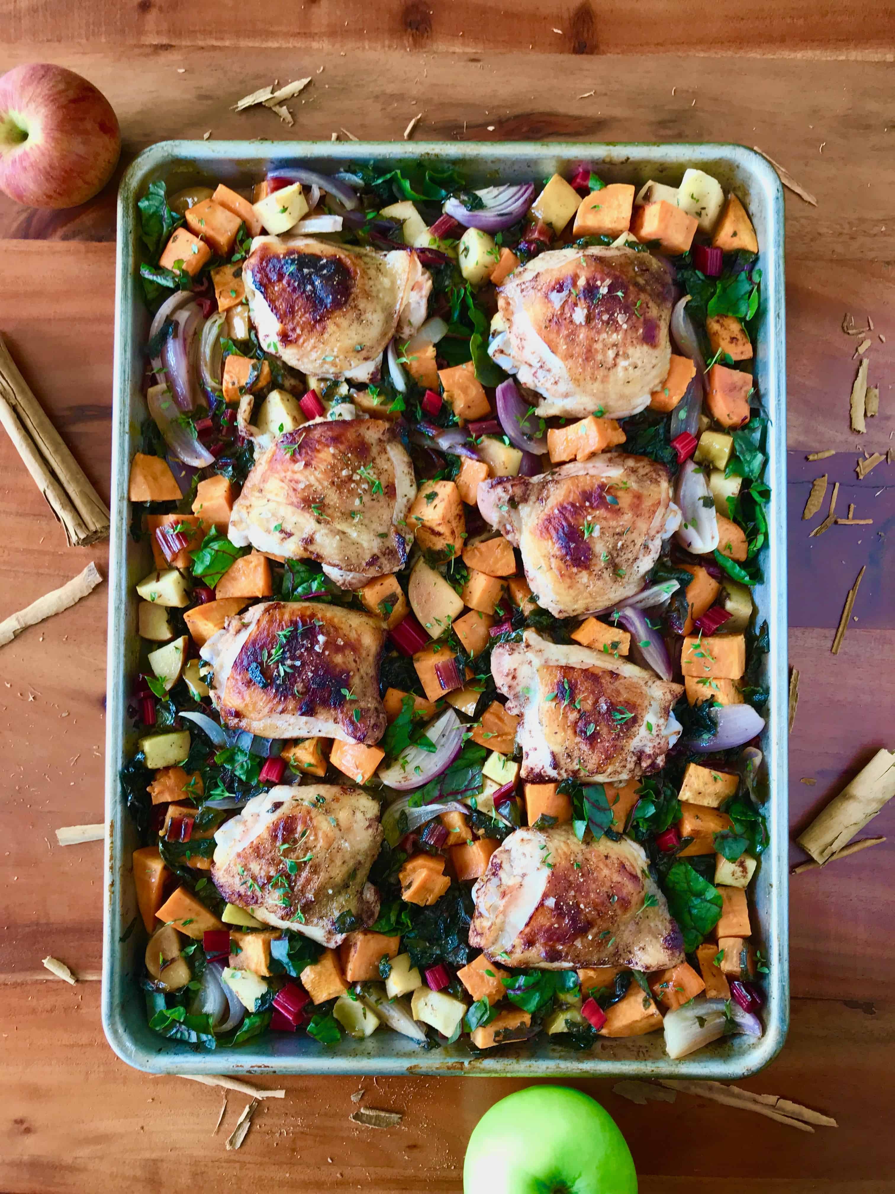 Sheet Pan Apple Cinnamon Chicken on a baking sheet on a wooden table surrounded by splintered cinnamon sticks and apples