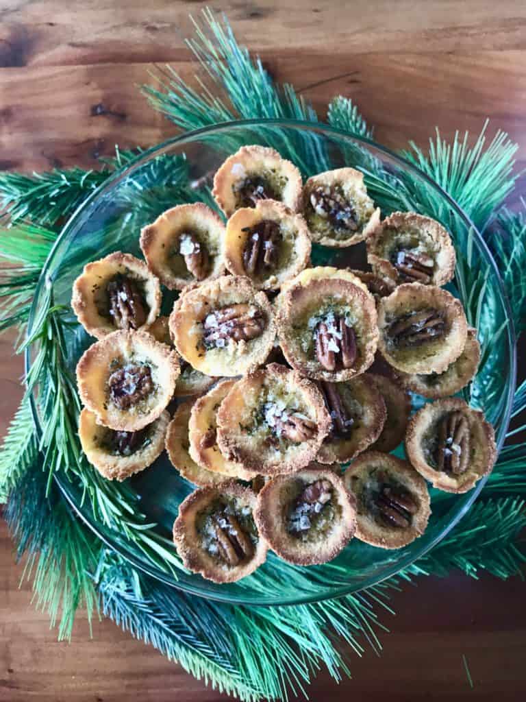 Mini paleo pecan pies on a glass cake stand surrounded by garland on a wooden table