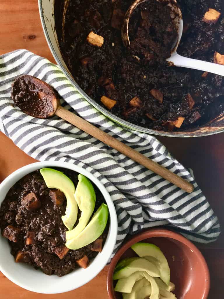Halloween Chili in a big green pot and a small white bowl with a striped kitchen towel, wooden spoon, ladle and small bowl of avocado slices