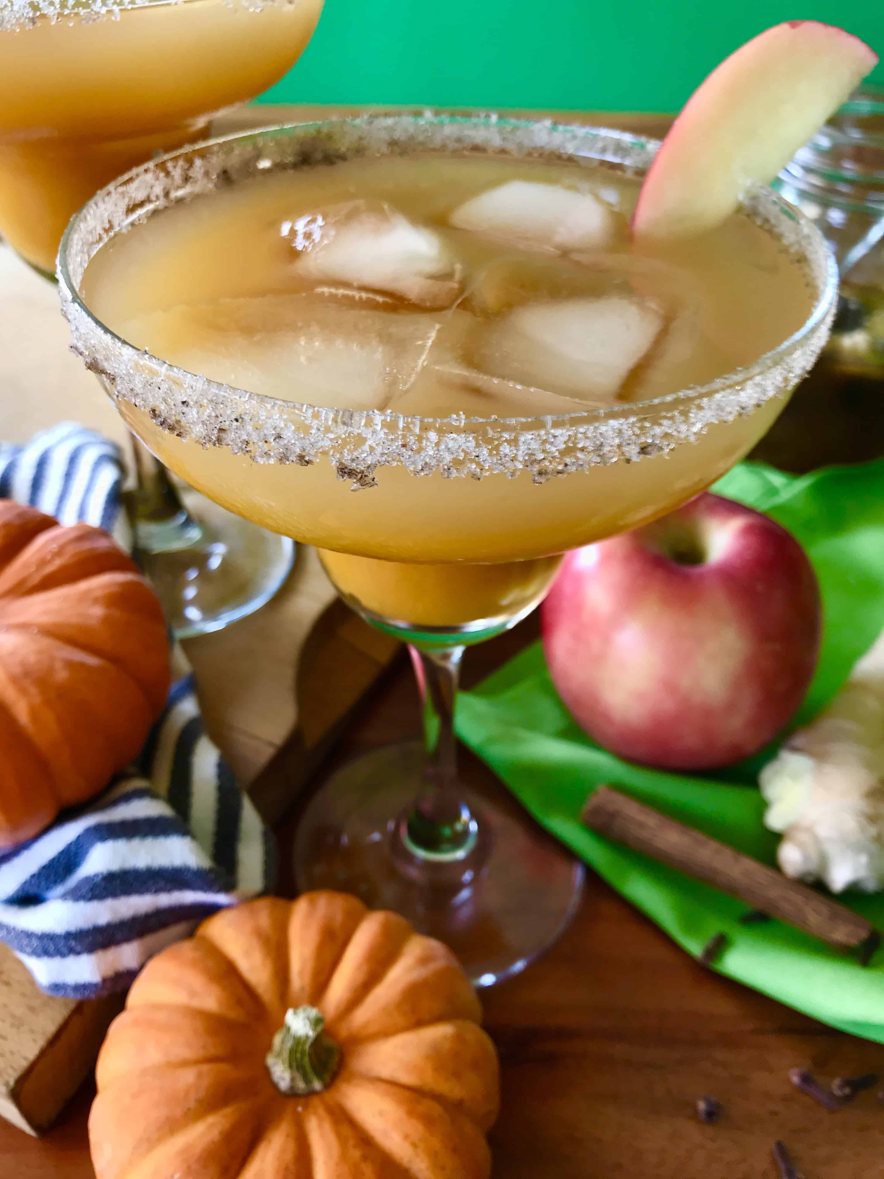 Pumpkin Cider Margarita on a wooden table surrounded by pumpkins, apples, cinnamon sticks, cloves, ginger, a striped towel, a green napkin and a jar of spiced simple syrup