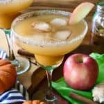 Pumpkin Cider Margarita on a wooden table surrounded by pumpkins, apples, cinnamon sticks, cloves, ginger, a striped towel, a green napkin and a jar of spiced simple syrup