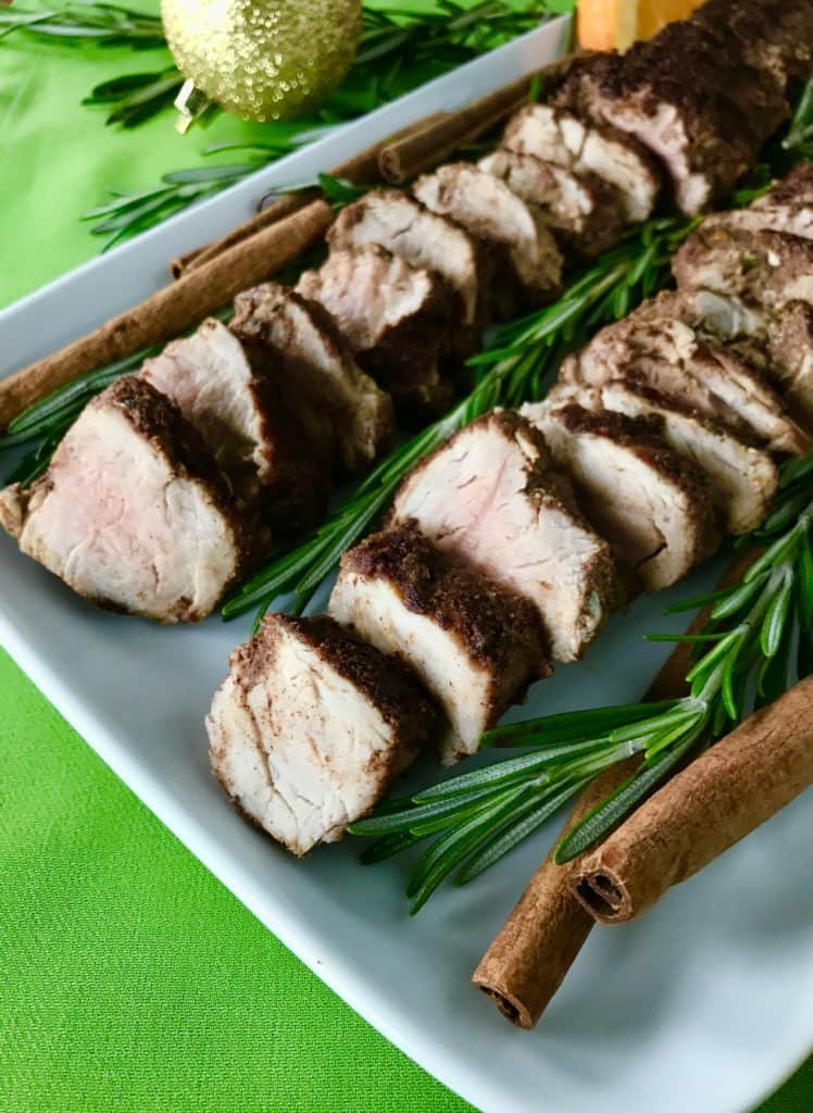 Christmas-Spiced Pork Tenderloin sliced on a white platter with rosemary, cinnamon sticks and orange wedges, on a green napkin