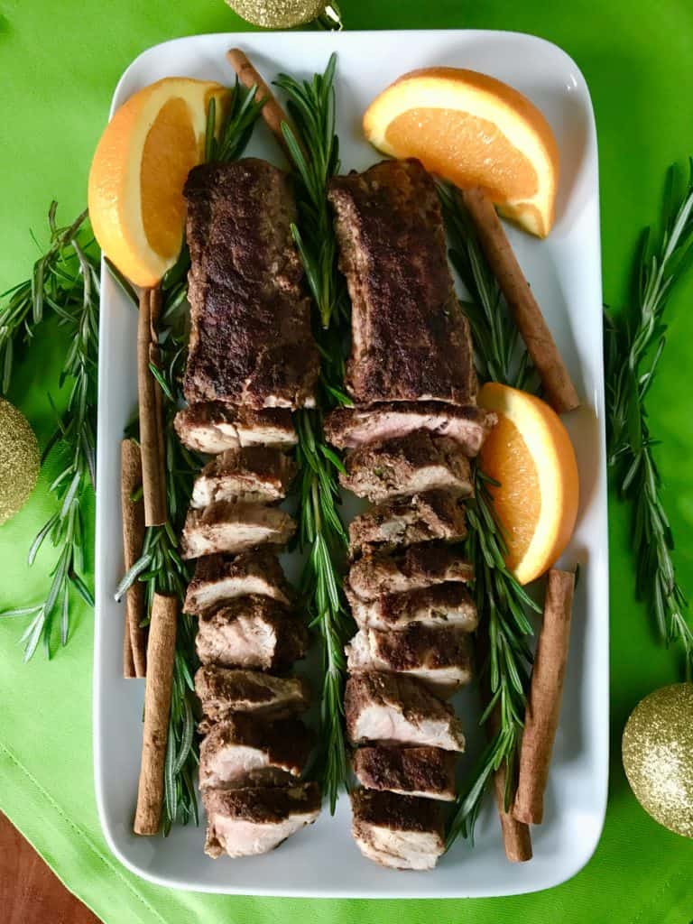 Christmas-Spiced Pork Tenderloin sliced on a white platter with rosemary, cinnamon sticks and orange wedges, on a green napkin with rosemary and shiny gold ornaments