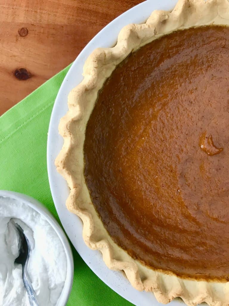 Paleo Pumpkin Pie next to a bowl of whipped coconut cream with a spoon in it, all on a green napkin on a wooden table