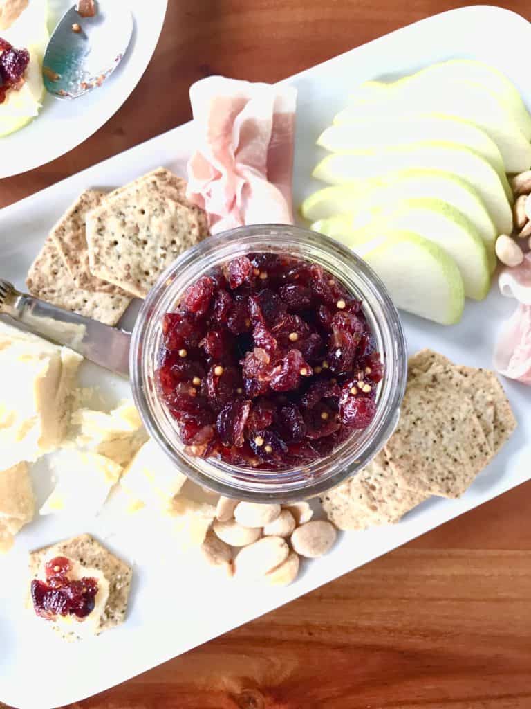 Savory, sweet, tart, and spicy Cranberry Prosecco Chutney served in a glass jar on a white platter with meat, cheese and crackers