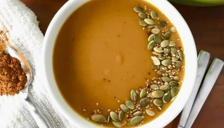 an image of curried sweet potato soup garnished with seeds in a white bowl next to a green pot with more soup, spoons and white towel, all on a wooden table