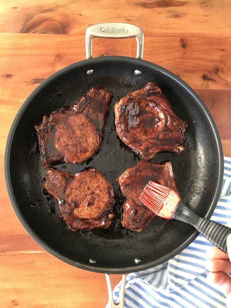 pan seared pork chops in a skillet getting brushed with a balsamic glaze, all on a wooden table with a blue striped towel next to it