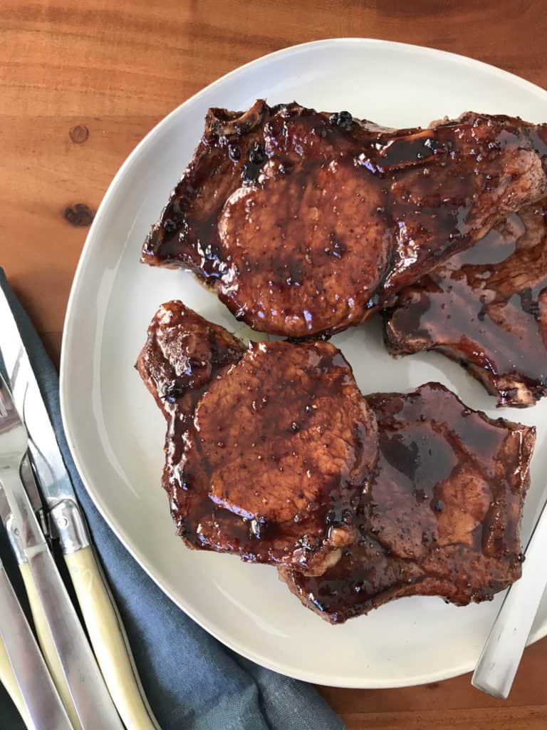 jammy balsamic pork chops on a white plate, next to a blue napkin and silverware, all on a wooden table