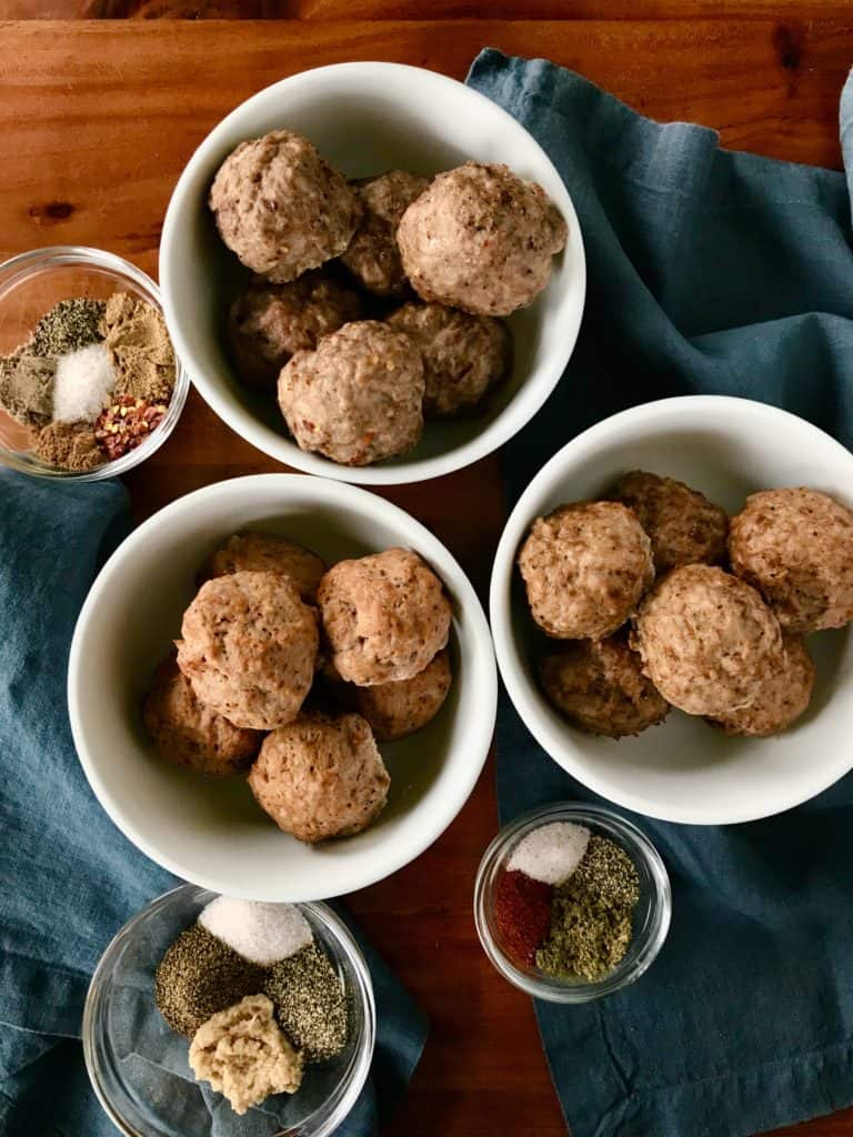 3 meatballs in white bowls next to blue napkins and little glass bowls of spices, all on a wooden table