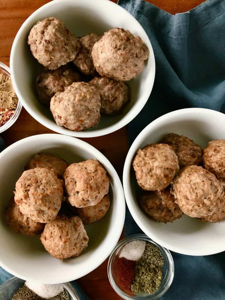 paleo meatballs in white bowls next to blue napkins and little glass bowls of spices, all on a wooden table