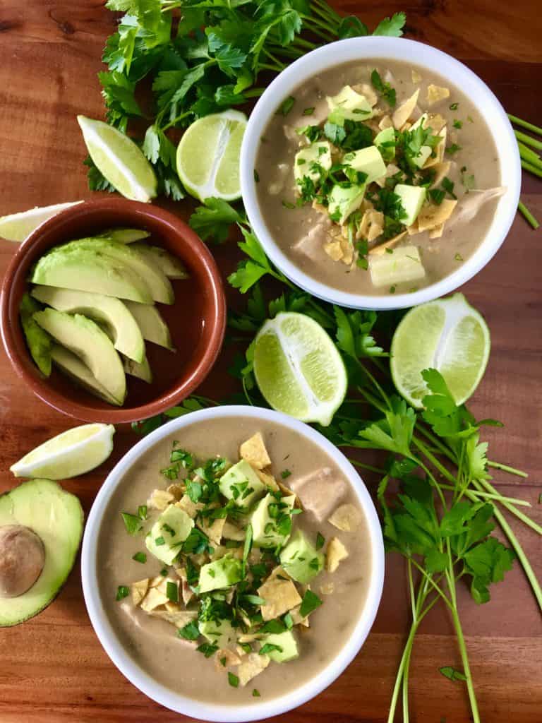creamy white chicken chili in white bowls on wooden tables surrounded by limes, avocado and parsley