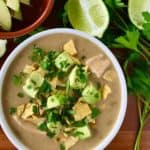 creamy white chicken chili in white bowls on wooden tables surrounded by limes, avocado and parsley