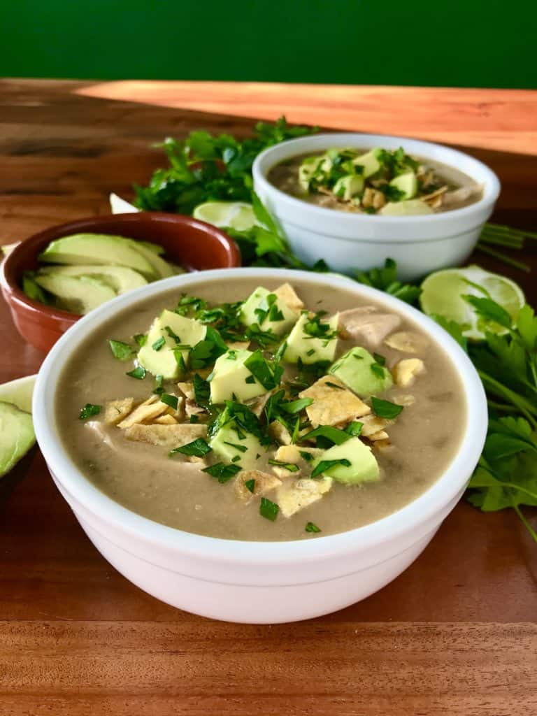 healthy white chicken chili in white bowls on wooden tables surrounded by limes, avocado and parsley