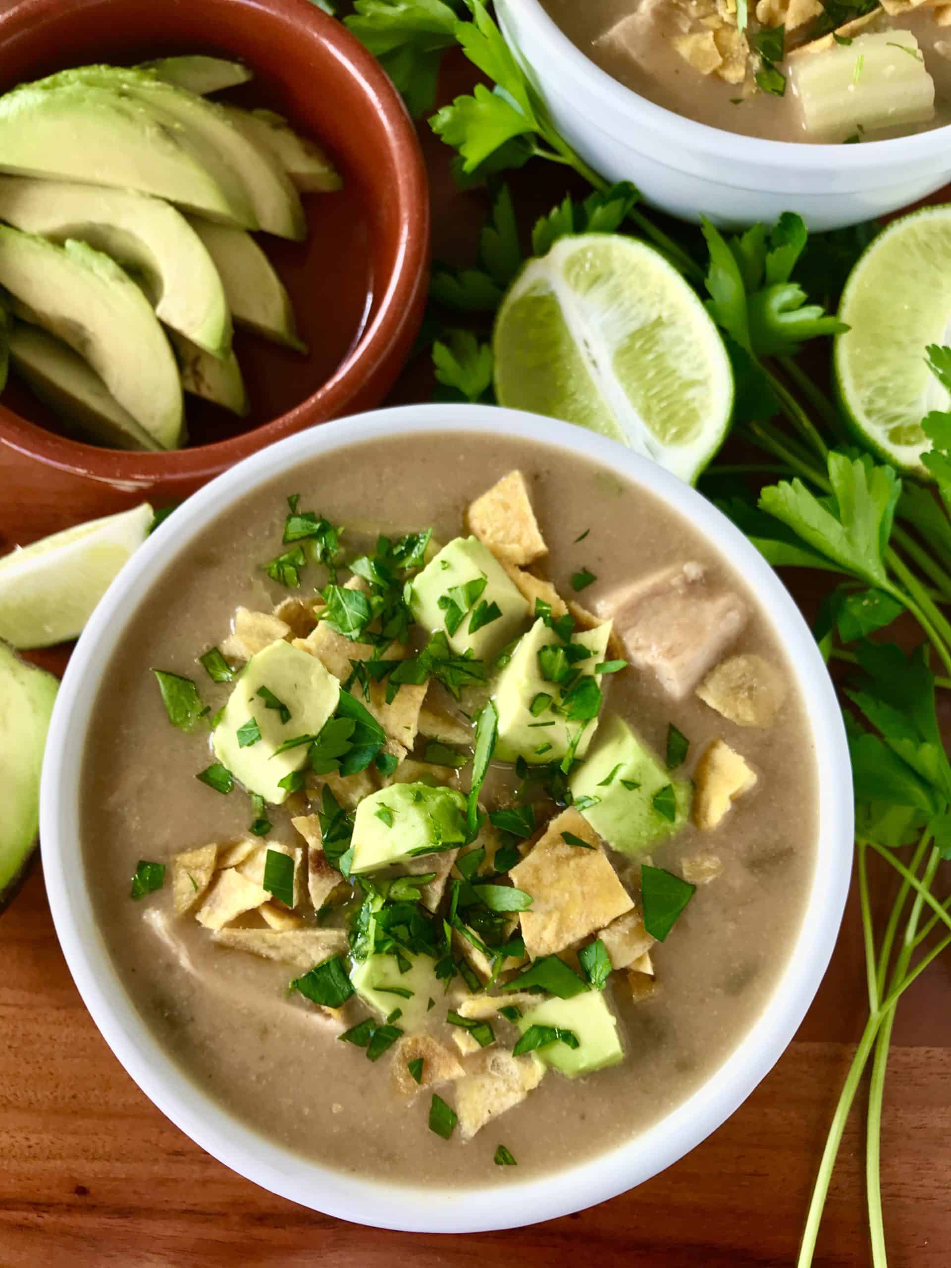 the best white chicken chili in white bowls on wooden tables surrounded by limes, avocado and parsley