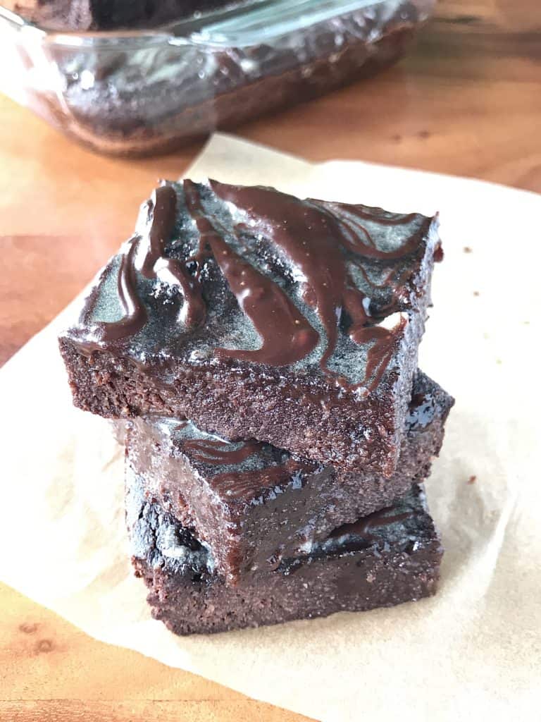 An image fudgy flourless brownies stacked on top of each other on parchment paper with the glass baking dish behind them, all on a wooden table