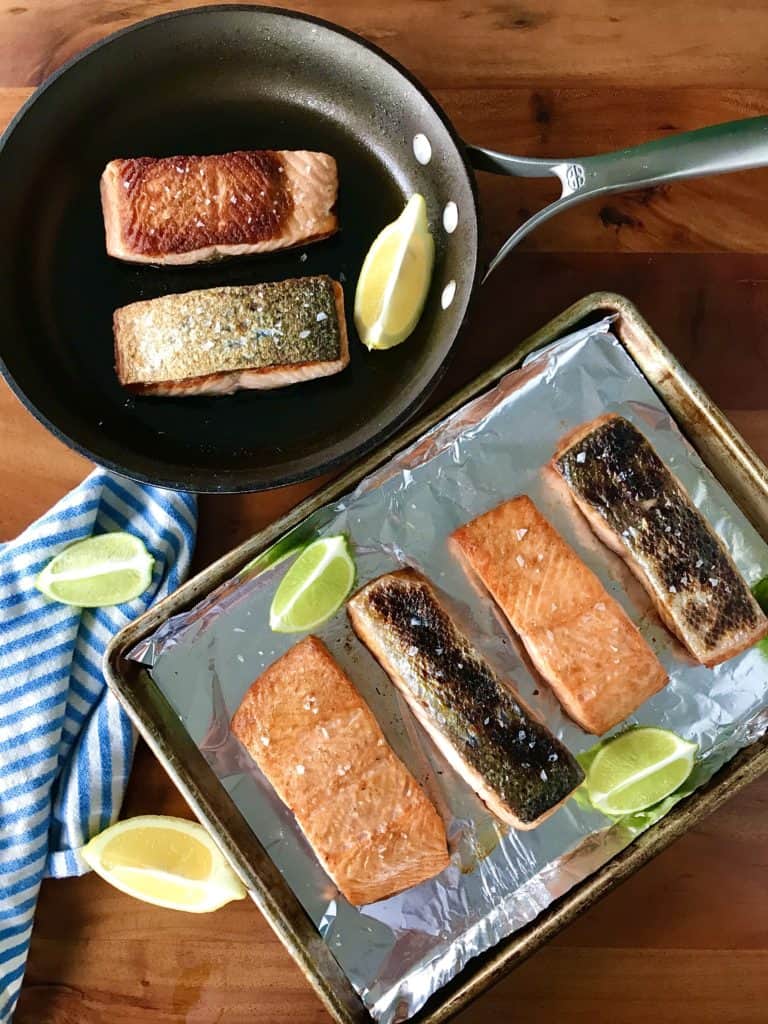 easy, healthy salmon is shown broiled on a baking sheet and pan seared in a skillet, both on a wooden table next to a blue striped towel and lemon and lime wedges