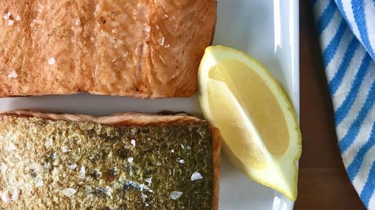 salmon with crispy skin on a white platter with a lemon and lime wedge, next to a blue and white striped towel, all on a wooden table