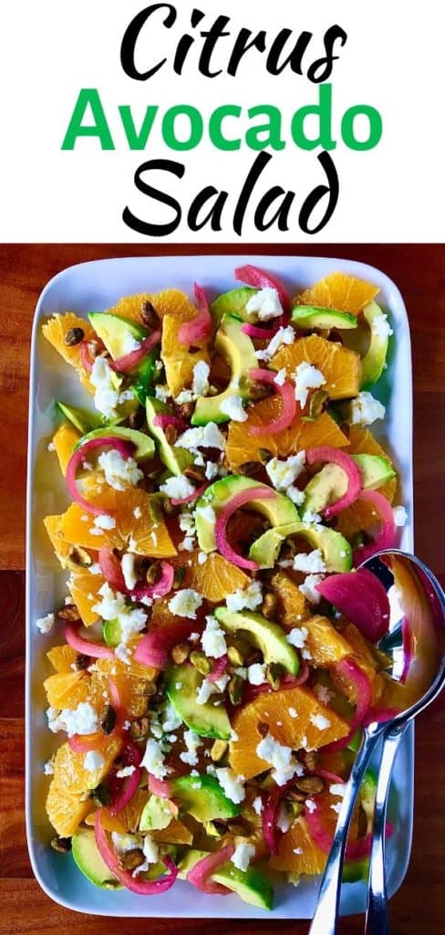 oranges, avocado, red onion, feta and pistachios on a white platter with stainless steel serving utensils on a wooden table