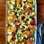 oranges, avocado, red onion, feta and pistachios on a white platter with stainless steel serving utensils on a wooden table