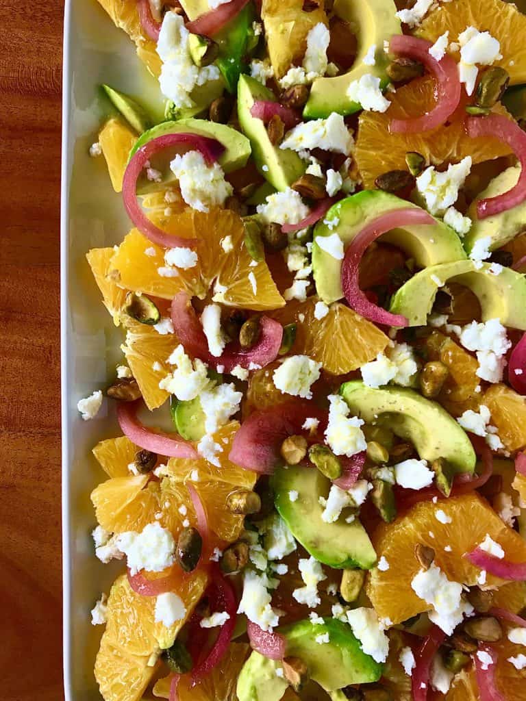 close up of oranges, avocado, onion, feta and pistachios on a white platter