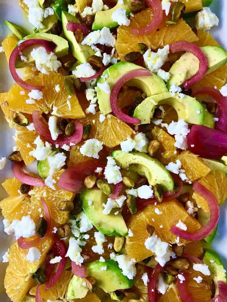 close up of oranges, avocado, onion, feta and pistachios on a white platter